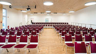 Foto eines mit roten Stühlen möblierten Hörsaals der Uni Witten. Man sieht eine große Holzdecke. 