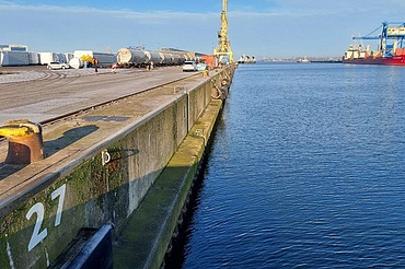 Picture shows a concrete quay in an industrial harbor, in the background a red ship