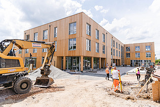 Foto der Uni Witten von außen. Man sieht ein großes Gebäude aus Holz und einen Bagger und Menschen.