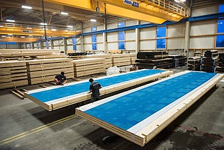 Foto in der Fertigung von ZÜBLIN Timber. Man sieht große Holzelemente mit einer blauen Wetterschutzfolie.