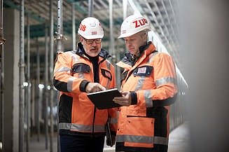 Zwei Arbeiter mit Sicherheitsbekleidung in einem Rohbau, die gemeinsam auf ein Tablet schauen