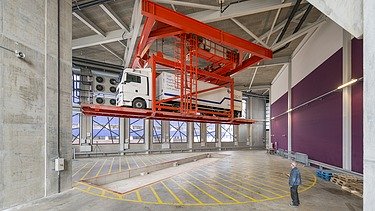 A grey hall with a red truck/car lift platform on which a white lorry is standing. An employee named Blau is standing at the front.