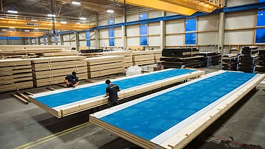 Foto in der Fertigung von ZÜBLIN Timber. Man sieht große Holzelemente mit einer blauen Wetterschutzfolie.