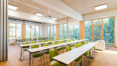 Klassenzimmer der Uni Witten. Man sieht einen möblierten Innenraum mit einer LENO-Holzdecke.