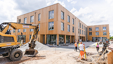 Foto der Uni Witten von außen. Man sieht ein großes Gebäude aus Holz und einen Bagger und Menschen.