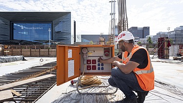 A picture of a building site; in the foreground, a man is kneeling next to an electrical box