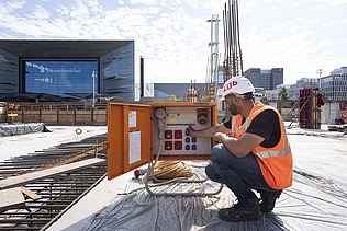 A picture of a building site; in the foreground, a man is kneeling next to an electrical box