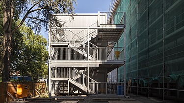 Picture of site huts and, on the right, the building covered in scaffolding with a green net