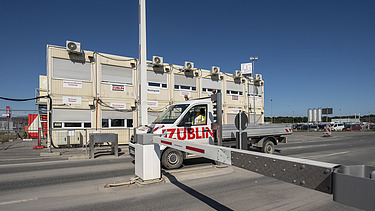 Bild von einer Zufahrtskontrolle, ein Auto fährt durch und Baucontainern im Hintergrund
