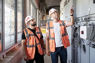 Foto mit zwei Mitarbeitern auf einer Baustelle vor Schränken mit der Haustechnik