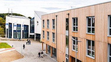 Foto der Uni Witten von außen. Man sieht ein großes Gebäudes aus Holz.