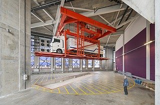 Eine graue Halle mit einer roten LKW/PKW Hebelbühne, auf der sich ein weißen Lastwagewn befindet. Vorne steht ein Mitarbeiter Blau.