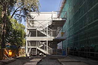 Bild von Baucontainern und rechts das Gebäude umhüllt von einem Baugerüst mit einem grünen Netz