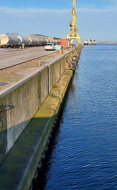Picture shows a concrete quay in an industrial harbor, in the background a red ship