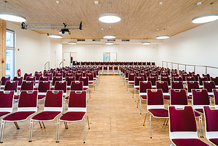 Foto eines mit roten Stühlen möblierten Hörsaals der Uni Witten. Man sieht eine große Holzdecke. 